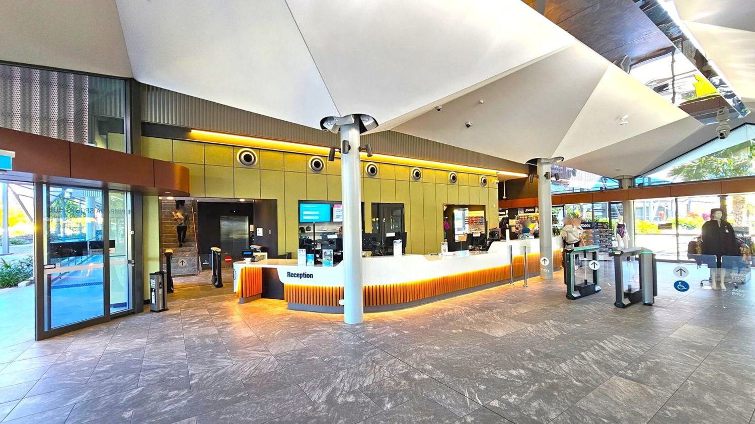 Reception area with a modern design. On the left, there are glass doors leading outside. In the centre, a white reception desk with orange lighting underneath, and several computer monitors. Behind the desk, a wall with circular vents and digital screens. On the right, people are entering through automated gates. The floor is tiled with a grey pattern, and the ceiling features large, angular white panels. Natural light streams in from large windows on the right.