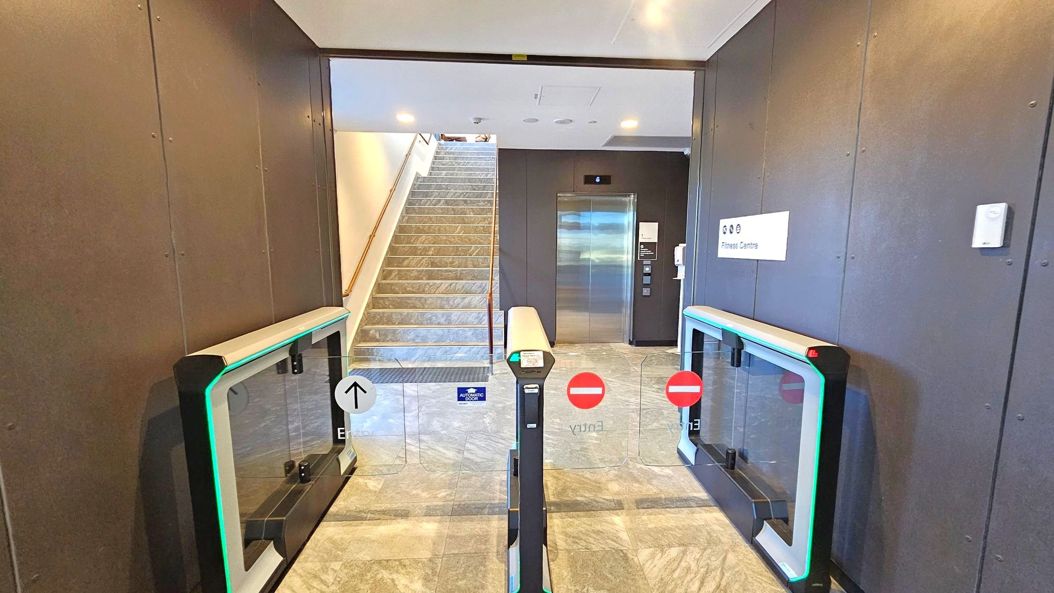 A modern entrance with electronic barriers featuring green lights and directional signs. In the centre, a staircase with a metal handrail leads upwards. To the right, a lift with a metallic door is visible, accompanied by a sign indicating a fitness centre. The floor is tiled with a grey pattern, and the ceiling is plain with recessed lighting. The walls are dark with a sleek finish.
