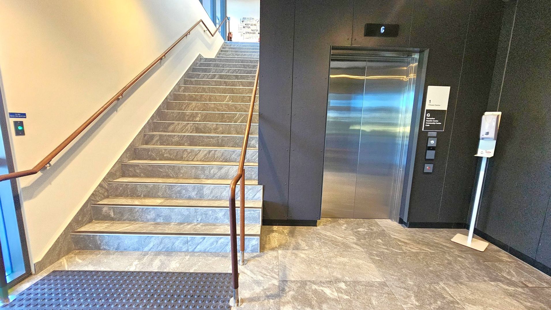 A stairway on the left with a wooden handrail leads upwards, featuring grey marble steps. On the right, a metallic lift with a digital display above shows the number 6. Next to the lift, a sign indicates floor information, and a hand sanitiser dispenser is mounted on a stand. The floor is covered with grey marble tiles, and a textured mat is placed at the base of the stairs. The walls are painted in a light colour, contrasting with the dark wall surrounding the lift.