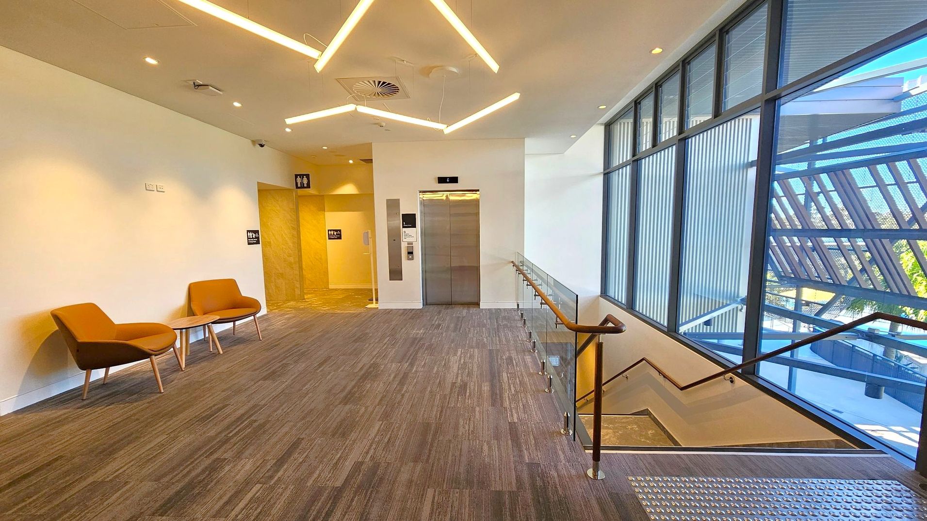 A modern interior space with a grey carpeted floor. On the left, two orange chairs with a small table are set against a white wall. In the centre, a lift with metallic doors is flanked by signs for restrooms. On the right, a staircase with a wooden handrail descends, next to large windows with vertical blinds, offering a view of an outdoor structure. The ceiling features geometric light fixtures and recessed lighting.