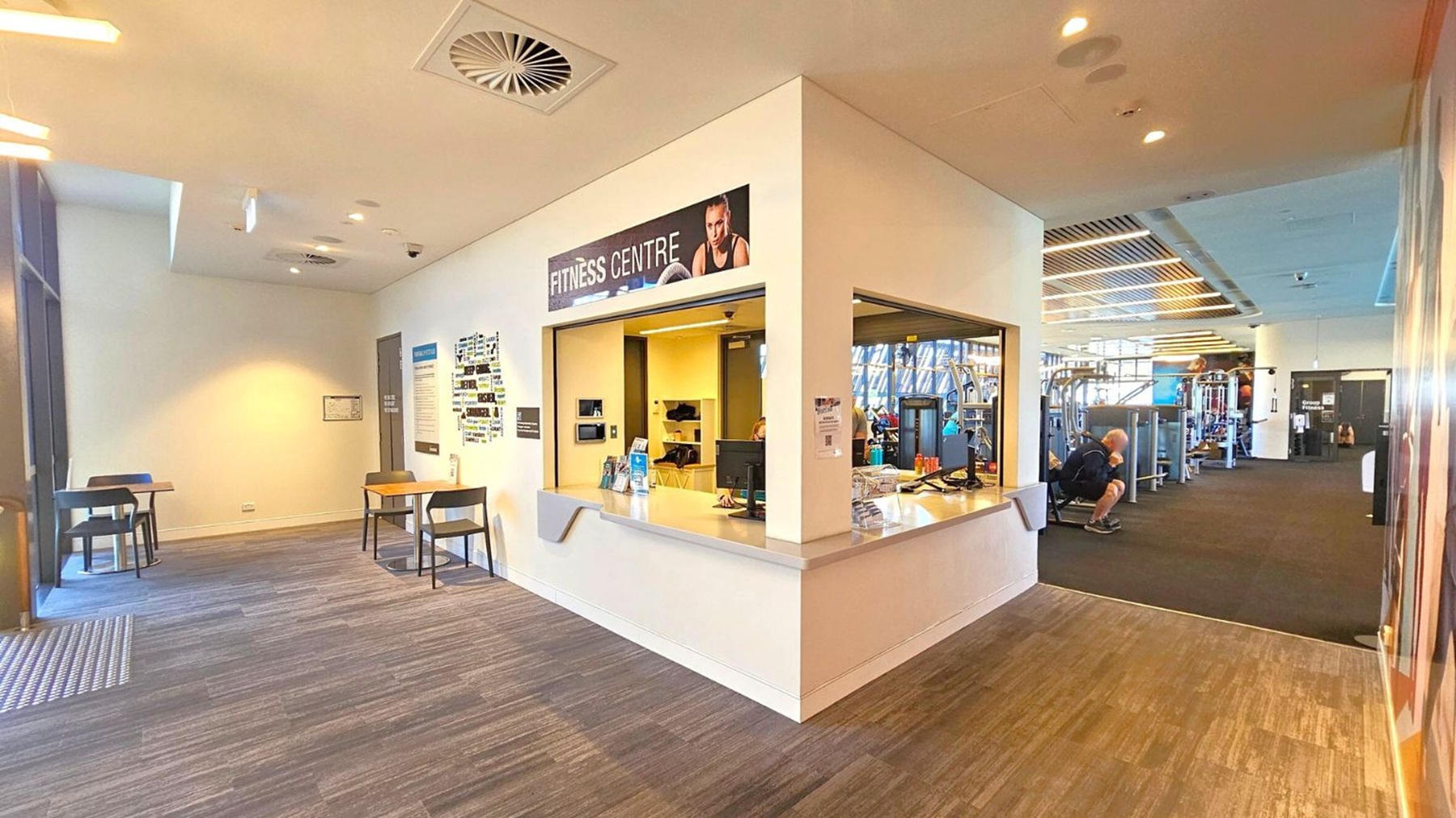 A fitness centre with a reception area in the middle, featuring a counter with a computer and water bottles. To the left, there are two small tables with chairs against a light-coloured wall. On the right, the gym area is visible with exercise equipment and a person sitting on a machine. The floor is wooden, and the ceiling has recessed lighting and ventilation panels.