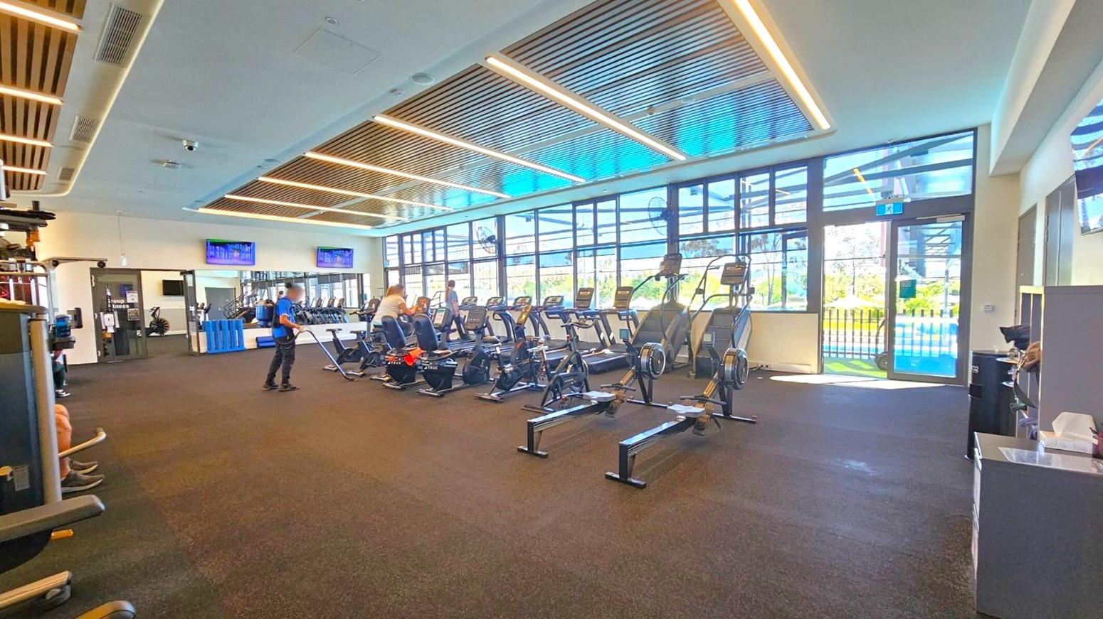 A spacious gym interior with a dark carpeted floor and a ceiling featuring wooden slats and recessed lighting. On the right, there are large windows allowing natural light to enter, with several treadmills and exercise machines lined up. In the centre, a person is standing near rowing machines. On the left, more gym equipment is visible. The background includes wall-mounted TVs and a reception area with a counter.