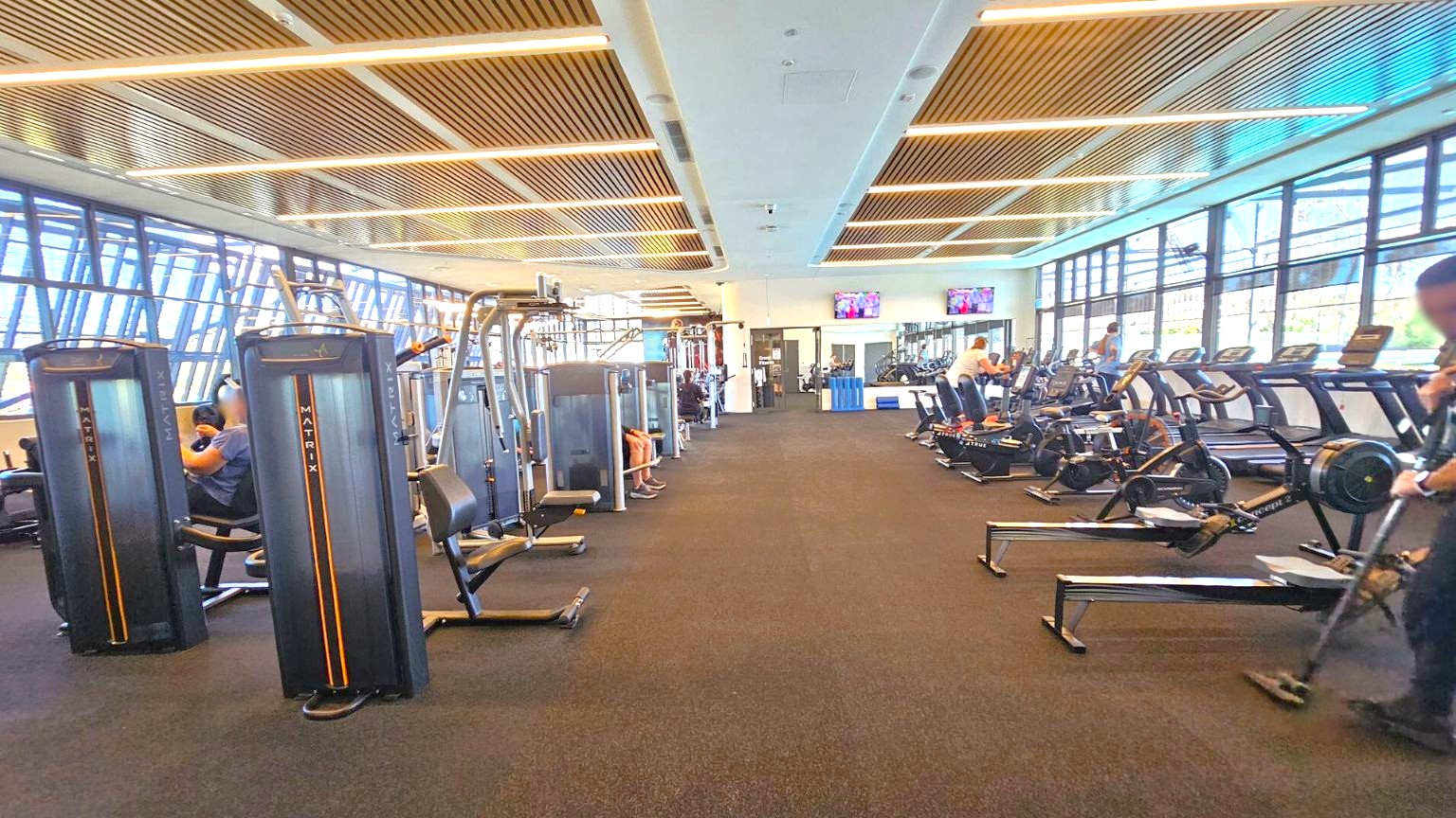 A spacious gym with a dark carpeted floor and a ceiling featuring wooden slats and recessed lighting. On the left, there are various weight machines with people using them. On the right, there are treadmills and rowing machines, also in use. Large windows line the right wall, allowing natural light to fill the space. In the background, two wall-mounted televisions are visible above a water cooler and a blue recycling bin.
