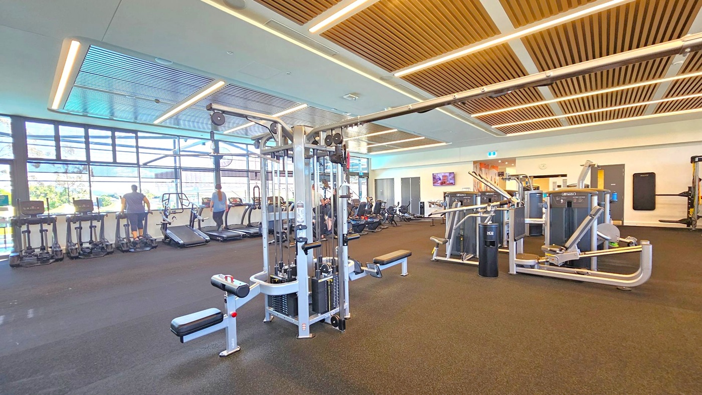A spacious gym with a variety of exercise equipment. On the left, there are several treadmills and elliptical machines near large windows that let in natural light. In the centre, a multi-station weight machine is visible. On the right, there are more weight machines and a wall-mounted television. The floor is covered with dark carpeting, and the ceiling features wooden slats with integrated lighting.