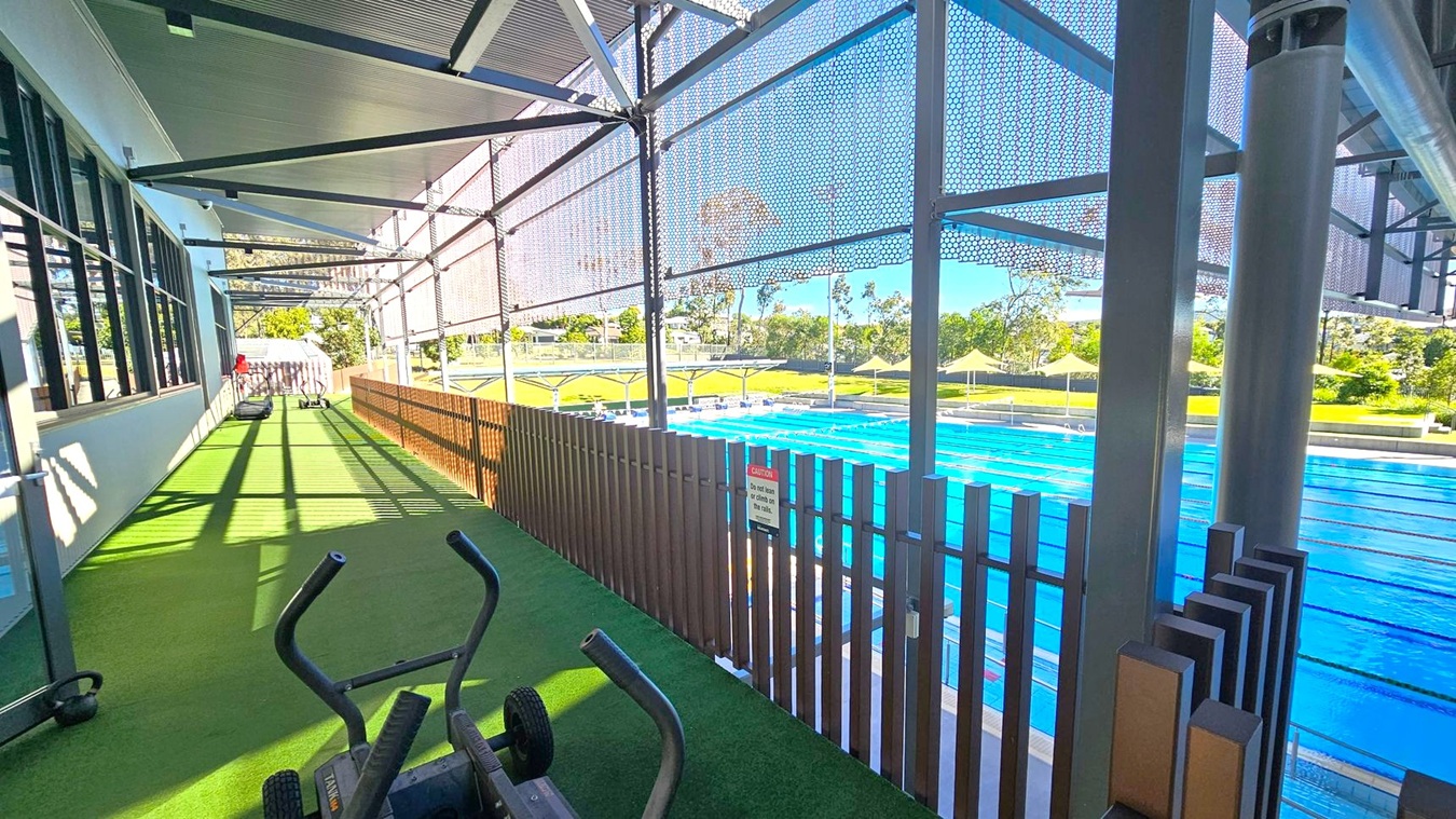 A gym area with green artificial turf flooring is on the left, featuring exercise equipment like stationary bikes. Large windows run along the left wall. On the right, a blue swimming pool is visible, separated by a wooden fence. The area is covered by a metal roof with a mesh screen, allowing sunlight to filter through. In the background, there are trees and a grassy area with shade structures.