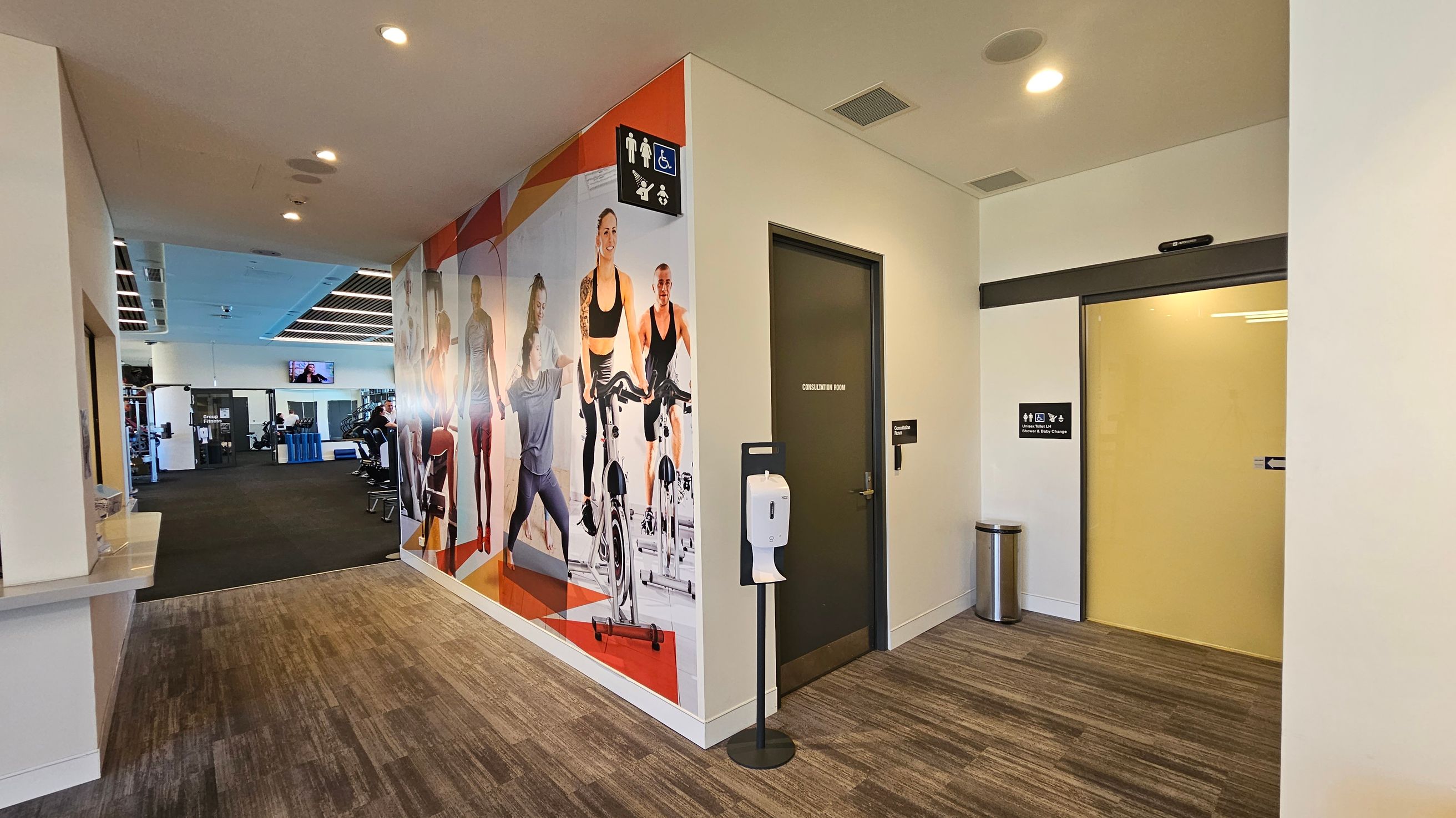 A gym interior with a corridor leading to a workout area on the left, featuring exercise equipment and a TV on the wall. The floor is carpeted in a mix of brown and grey tones. On the right, there is a wall with a large mural of people exercising, a door labelled "Consultation Room," and a sanitiser dispenser. A sign indicates accessible toilets and baby change facilities. The ceiling has recessed lighting.
