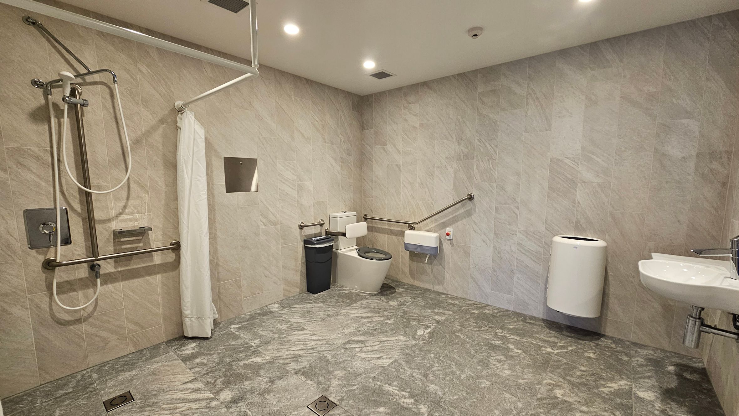 A spacious, accessible bathroom with light grey tiled walls and floor. On the left, there's a shower area with a handheld showerhead, grab bars, and a curtain. In the centre, a toilet with a black bin beside it and a grab bar on the wall. On the right, a white sink with a modern tap and a wall-mounted hand dryer. The ceiling is fitted with recessed lighting.