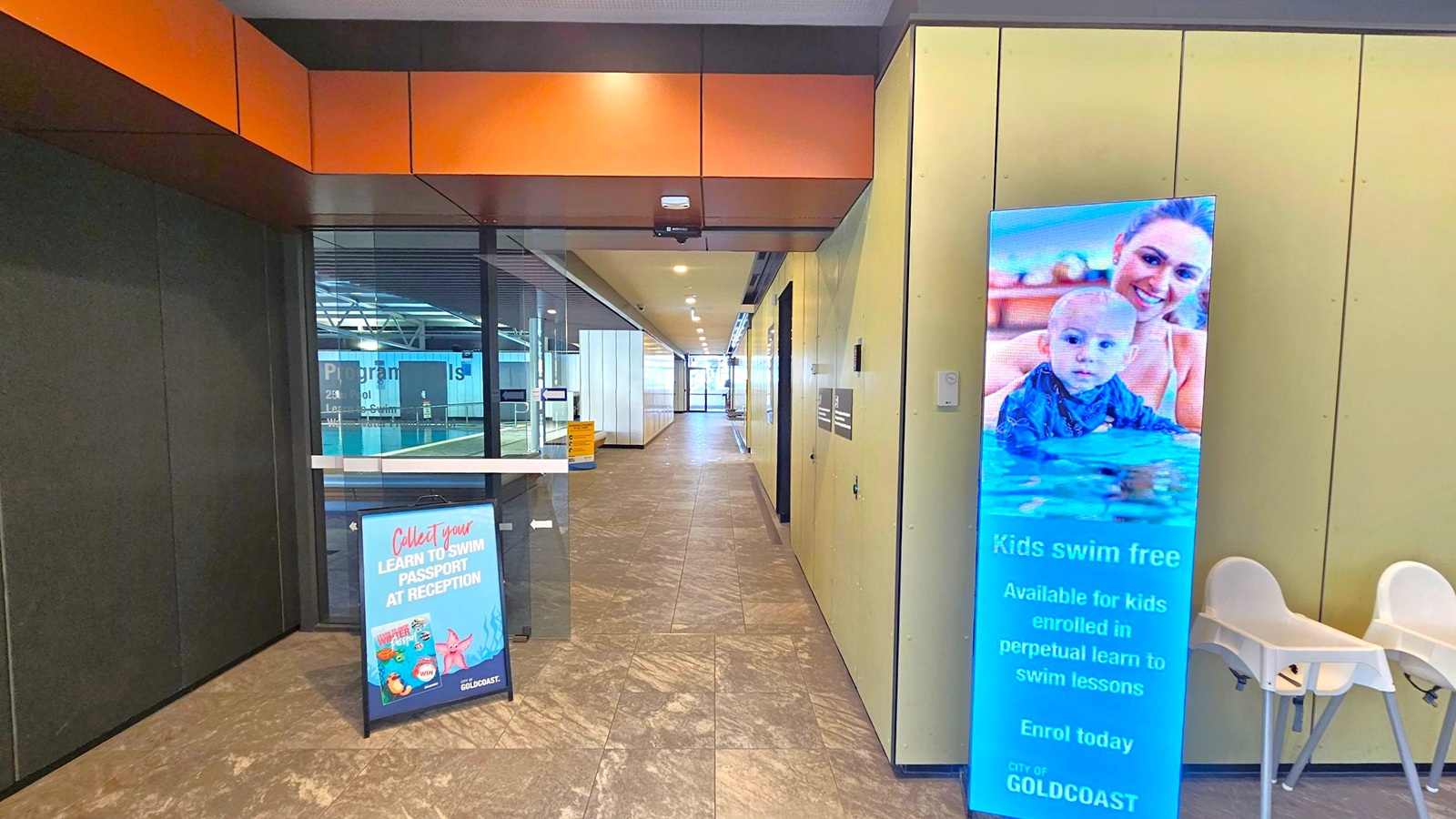 A hallway with a tiled floor and a ceiling with recessed lighting. On the right, there's a digital sign displaying an advertisement for free swimming lessons for kids, featuring an image of a woman and a baby in a pool. Next to the sign are two white high chairs. On the left, a stand with a sign promoting a "Learn to Swim Passport" at reception. The background shows glass doors leading to an indoor pool area.