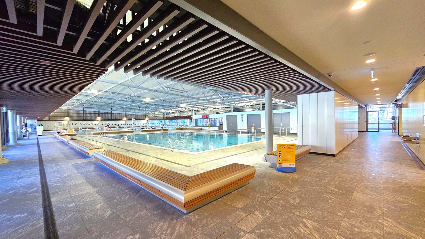 An indoor swimming pool area with a large pool in the centre. The floor is tiled with a grey pattern. Wooden benches line the pool's perimeter. The ceiling features a grid of beams and recessed lighting. On the right, a corridor leads to a glass exit. A sign on a stand near the pool provides safety instructions. The pool area is well-lit, and the walls have a combination of glass and solid panels.