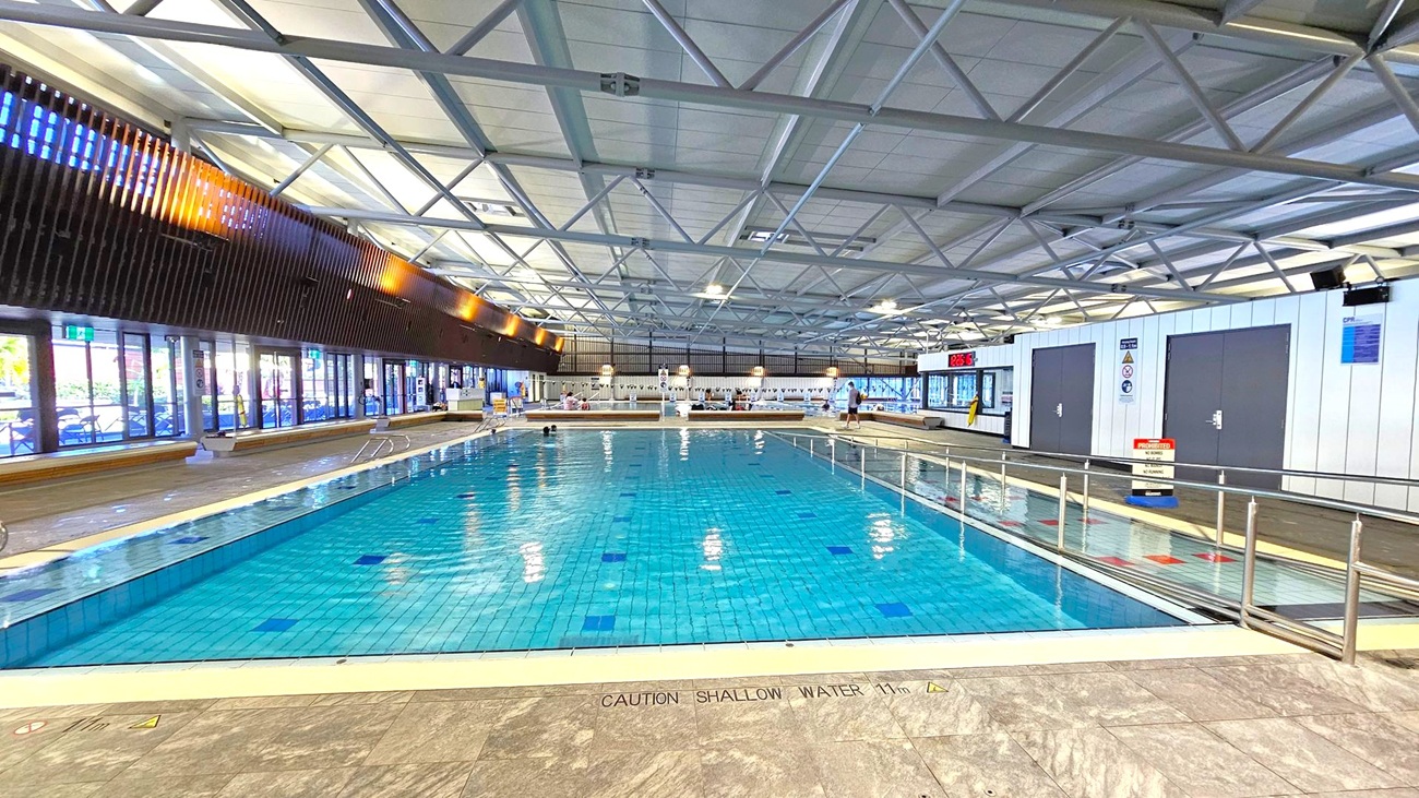 Indoor swimming pool with clear blue water, surrounded by a tiled deck. On the left, large glass doors and windows allow natural light in, with wooden slats above. The right side features white walls with dark doors and a digital clock displaying "23.6°C". The ceiling has a grid of metal beams and panels. A sign on the floor reads "CAUTION SHALLOW WATER 1.1m". A few people are visible near the pool's edge.