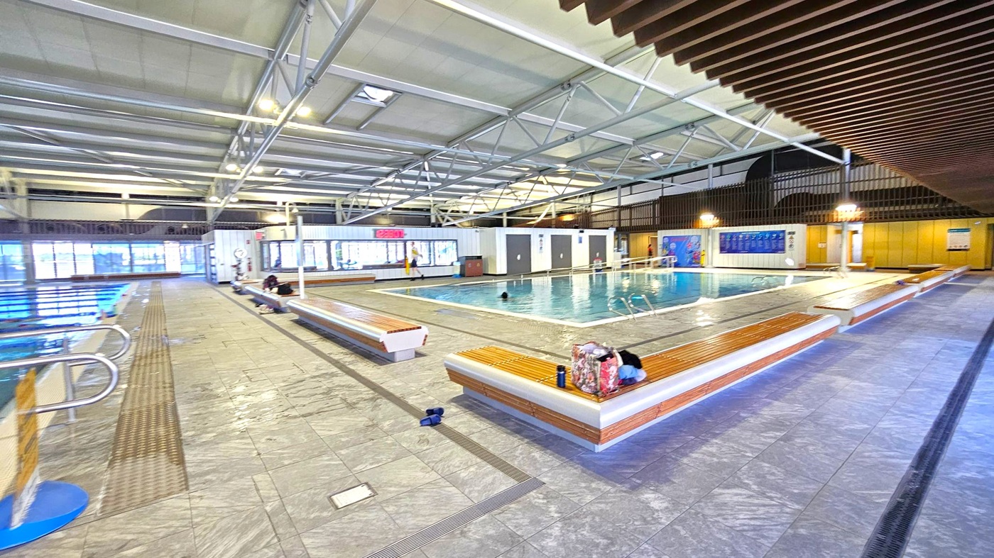 Indoor swimming pool area with a large pool in the centre. Benches with wooden slats surround the pool, with some personal items placed on them. The floor is tiled in grey. On the left, a smaller pool with metal handrails. The ceiling is high with exposed metal beams and fluorescent lighting. Large windows line the back wall, allowing natural light. A digital clock is mounted on the wall above the windows. The right side has a wooden slatted ceiling feature.