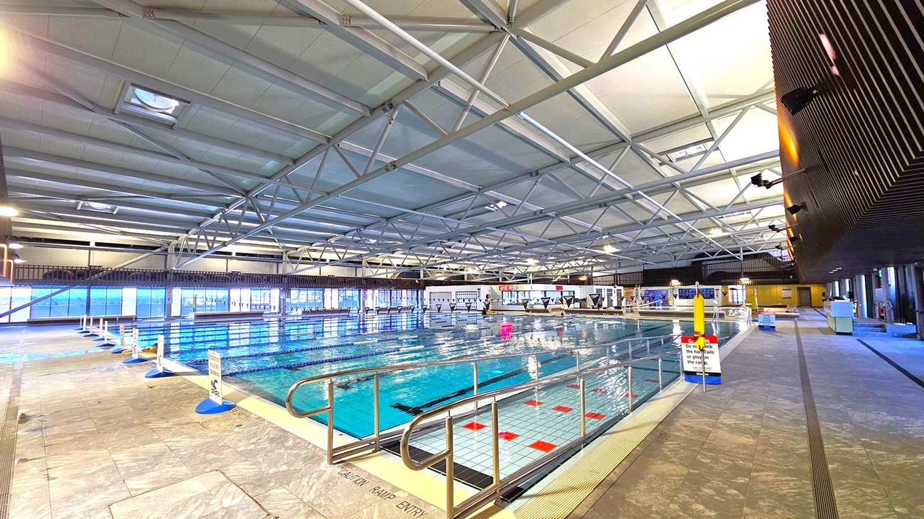 An indoor swimming pool with multiple lanes, surrounded by a tiled deck. On the left, large windows allow natural light to enter. The ceiling is high with a metal truss structure. On the right, there is a dark wall with lights and a walkway. Safety signs and pool equipment are visible around the pool area. The floor is tiled, and the pool has a ramp entry with handrails.