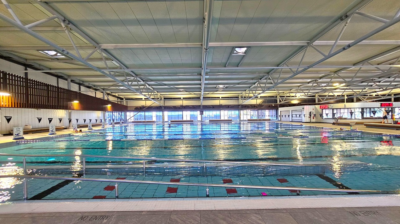An indoor swimming pool with clear water, divided into lanes by floating ropes. The ceiling features exposed metal beams and overhead lights. On the left, a series of triangular flags hang above the pool, and a wooden slatted wall runs along the side. The right side has digital clocks displaying times and a few people near the pool's edge. Large windows in the background allow natural light to illuminate the space. The floor is tiled with a "NO ENTRY" sign visible near the pool's edge.