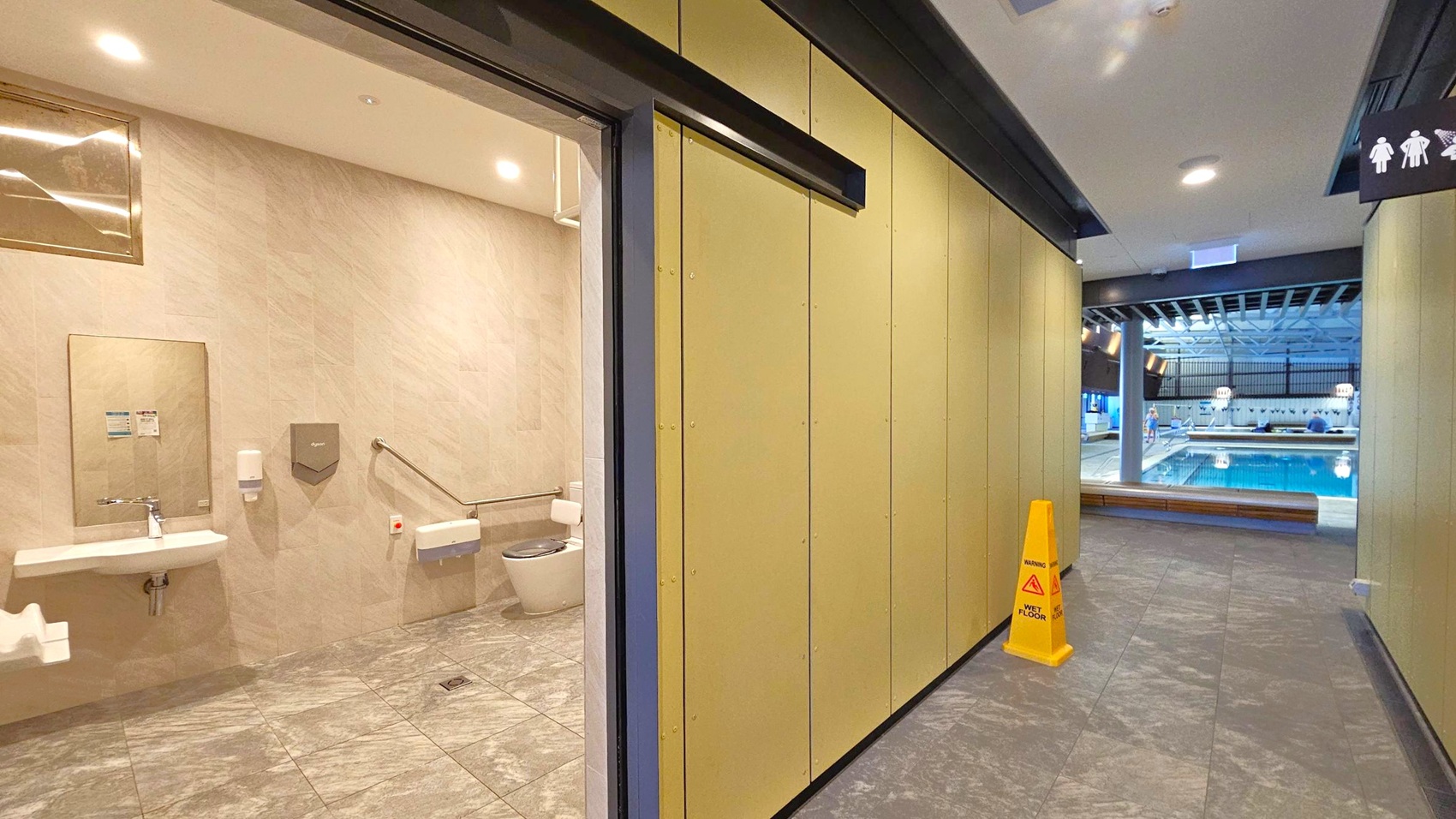 On the left, there's a restroom with a sink, mirror, and toilet equipped with handrails. The floor is tiled in a grey pattern. On the right, a corridor leads to a swimming pool area visible in the background. A yellow "Wet Floor" sign is placed on the corridor's tiled floor. The ceiling has recessed lighting, and a sign indicating restroom facilities is visible above the corridor.