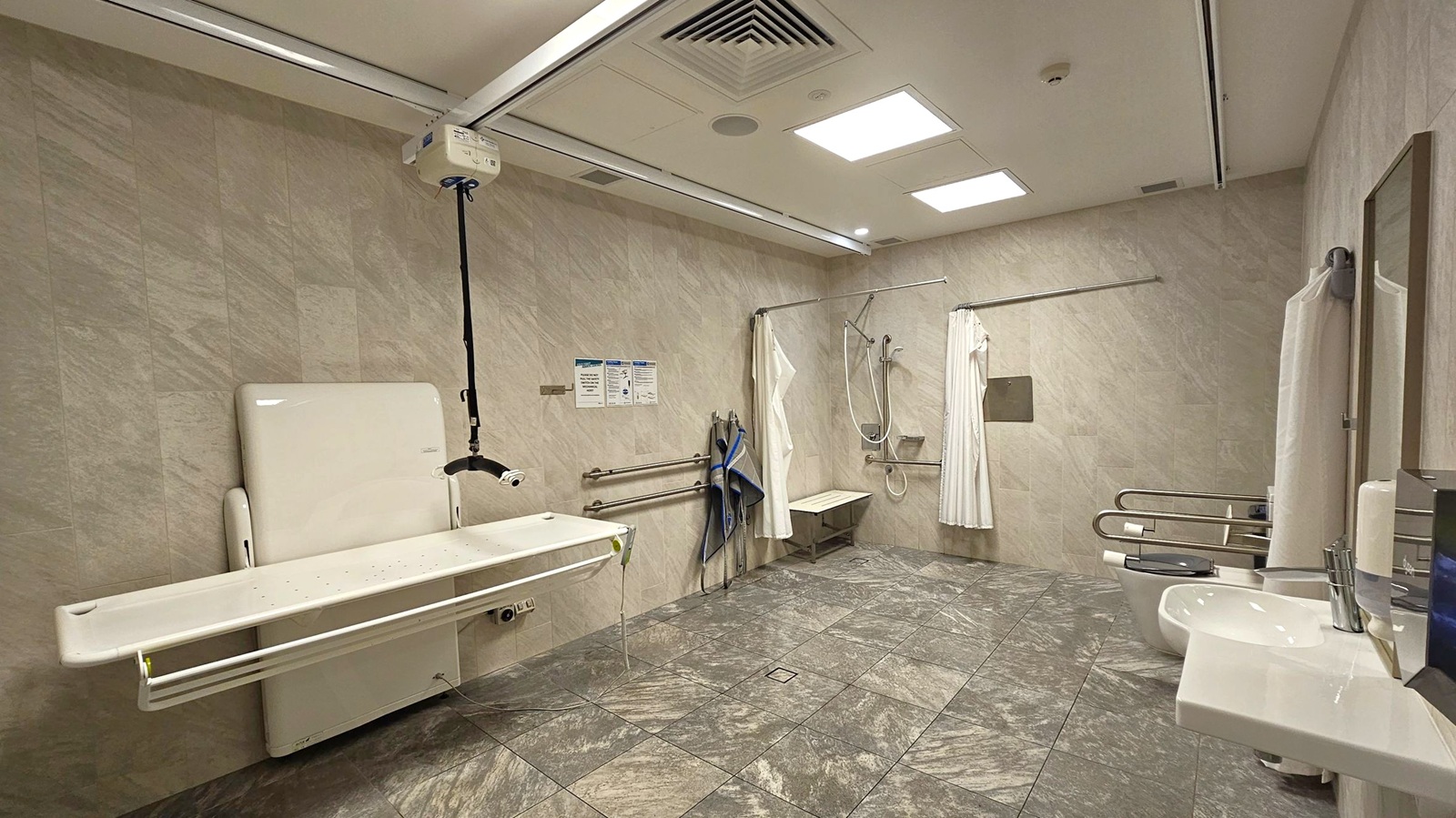 A spacious accessible bathroom with a green marbled floor and light-coloured walls. On the left, a foldable changing table is mounted on the wall with an overhead hoist system. In the centre, a shower area with a bench, grab bars, and a curtain. On the right, a sink with a mirror and a toilet equipped with support rails. The ceiling has recessed lighting and ventilation panels. Towels hang on the wall near the shower area.