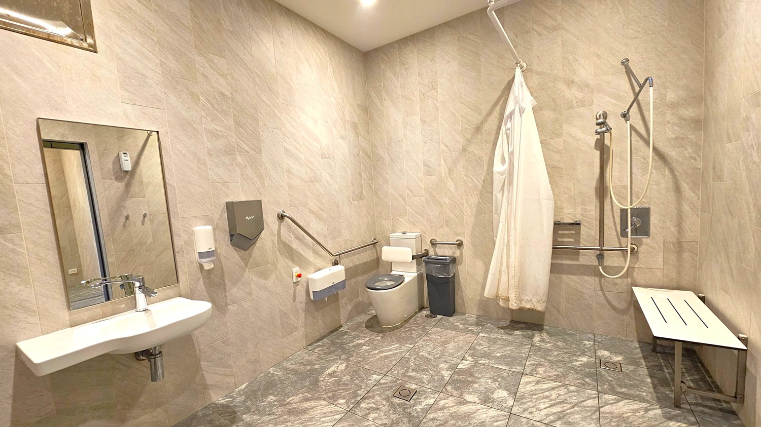 Accessible bathroom with grey tiled walls and floor. On the left, a wall-mounted sink with a mirror above, a soap dispenser, and a hand dryer. In the centre, a toilet with support rails and a small bin beside it. On the right, a shower area with a curtain, handheld showerhead, and a foldable bench. The ceiling has recessed lighting.