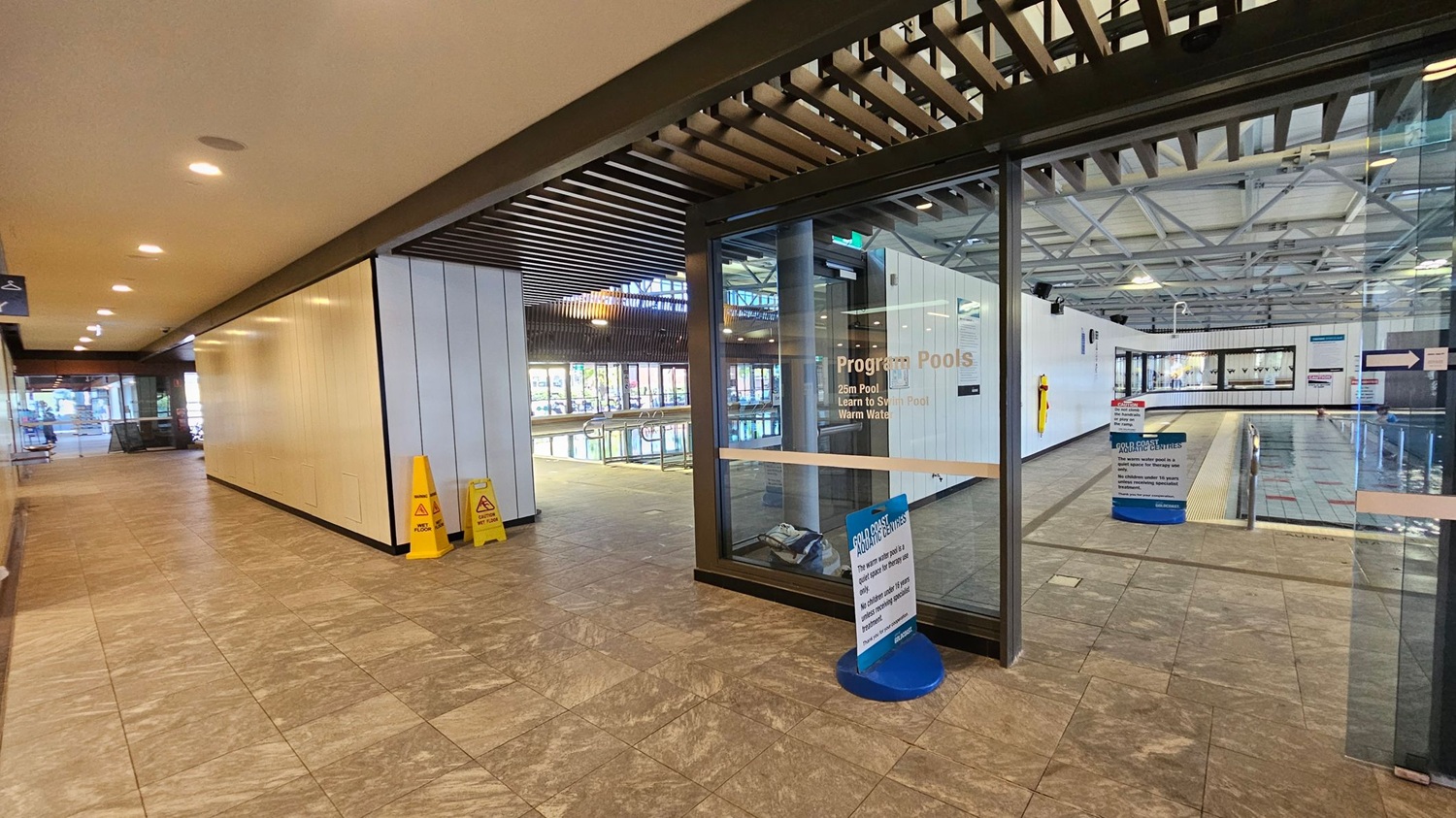 A corridor with a tiled floor leads to a glass entrance on the right, marked "Program Pools." Inside, a swimming pool is visible with a tiled deck. Two yellow caution signs are placed near the entrance. The ceiling features recessed lighting and a slatted design. On the left, the corridor extends with more lighting and a bench in the distance. The pool area has large windows and a high, industrial-style ceiling with exposed beams.