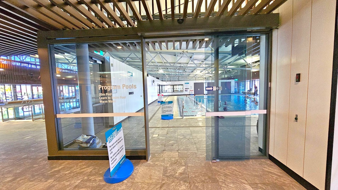 Glass doors lead to an indoor swimming pool area. The left side shows a pool with people swimming, visible through large windows. The right side has a wall with a control panel. In the centre, a sign on a stand provides information about pool rules. The floor is tiled, and the ceiling features exposed beams and lighting. The pool area is well-lit with natural light coming through the windows.