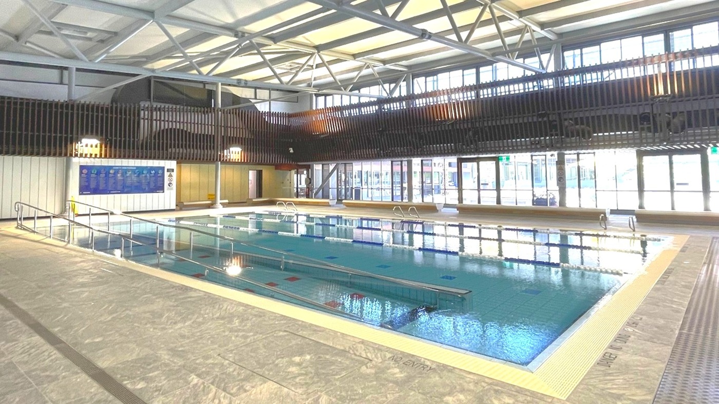 Indoor swimming pool with clear water, surrounded by a tiled deck. On the left, a metal handrail leads into the pool. The background features large windows allowing natural light, and a wooden slatted wall above. The ceiling has exposed metal beams and skylights. A blue information board is visible on the left wall.