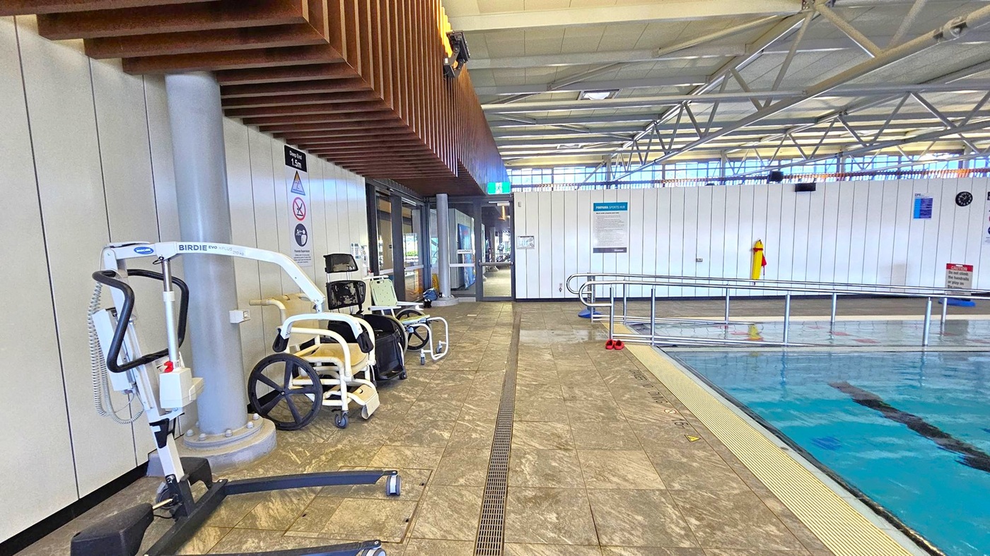 On the left, there is a pool lift and a wheelchair near a column, with a wooden slatted ceiling above. In the middle, a corridor leads to a glass door, with signs and a green exit sign visible. On the right, a swimming pool with a metal railing along the edge. The floor is tiled with a drainage grate running along it. The ceiling has exposed metal beams and large windows letting in natural light.