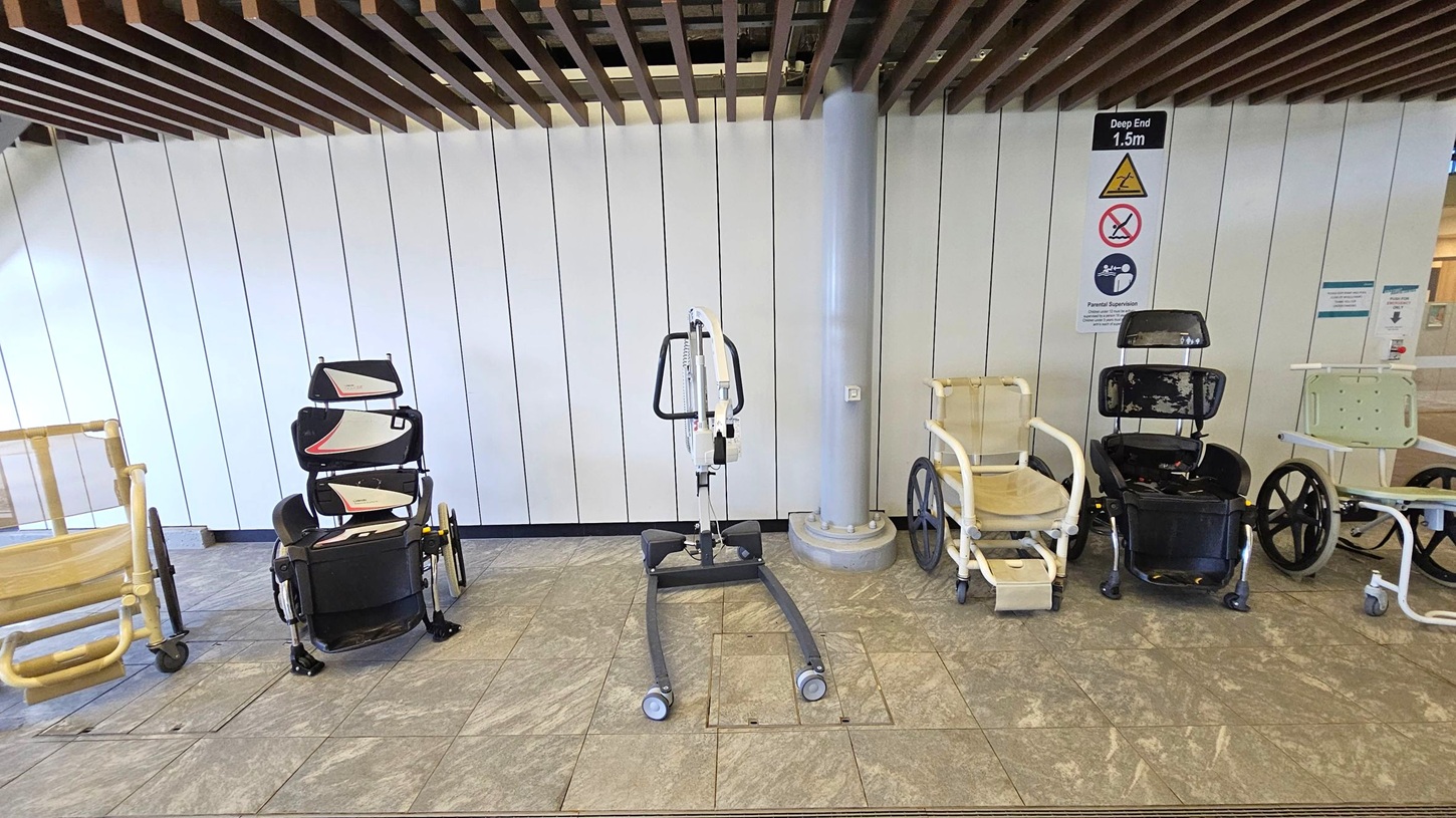 A row of mobility aids is lined up against a white panelled wall. On the left, there are two beige wheelchairs with large wheels. In the centre, a mechanical lift with a metal frame and wheels. To the right, two black wheelchairs with high backs and a white shower chair with wheels. The floor is tiled in a light brown pattern, and the ceiling features wooden slats. Signs on the wall indicate a deep end of 1.5 metres and other safety instructions.