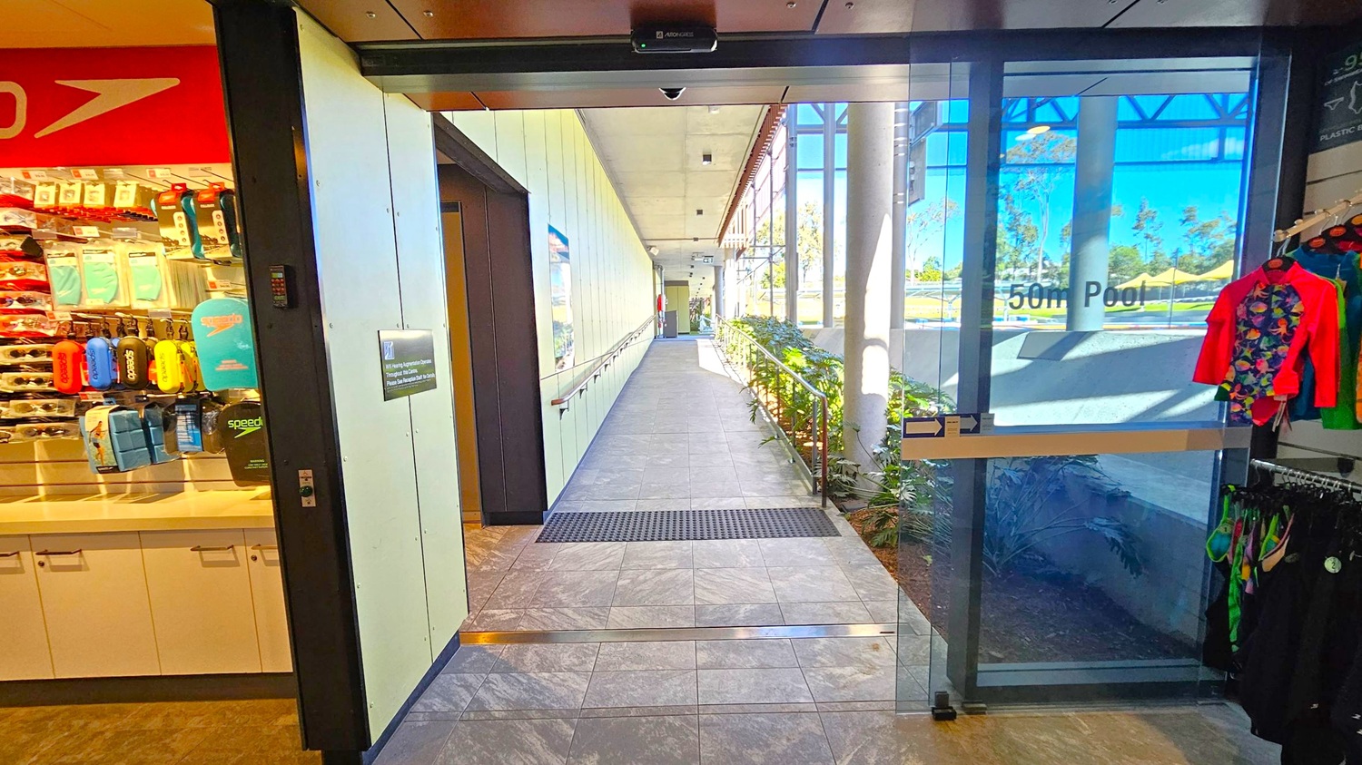 A corridor with a tiled floor leads to a glass door marked "50m Pool" on the right. On the left, there's a shop area displaying swimming gear, including goggles and colourful swimwear. The corridor has a handrail and a row of plants along the right side. The ceiling is fitted with lights, and large windows allow natural light to illuminate the space.