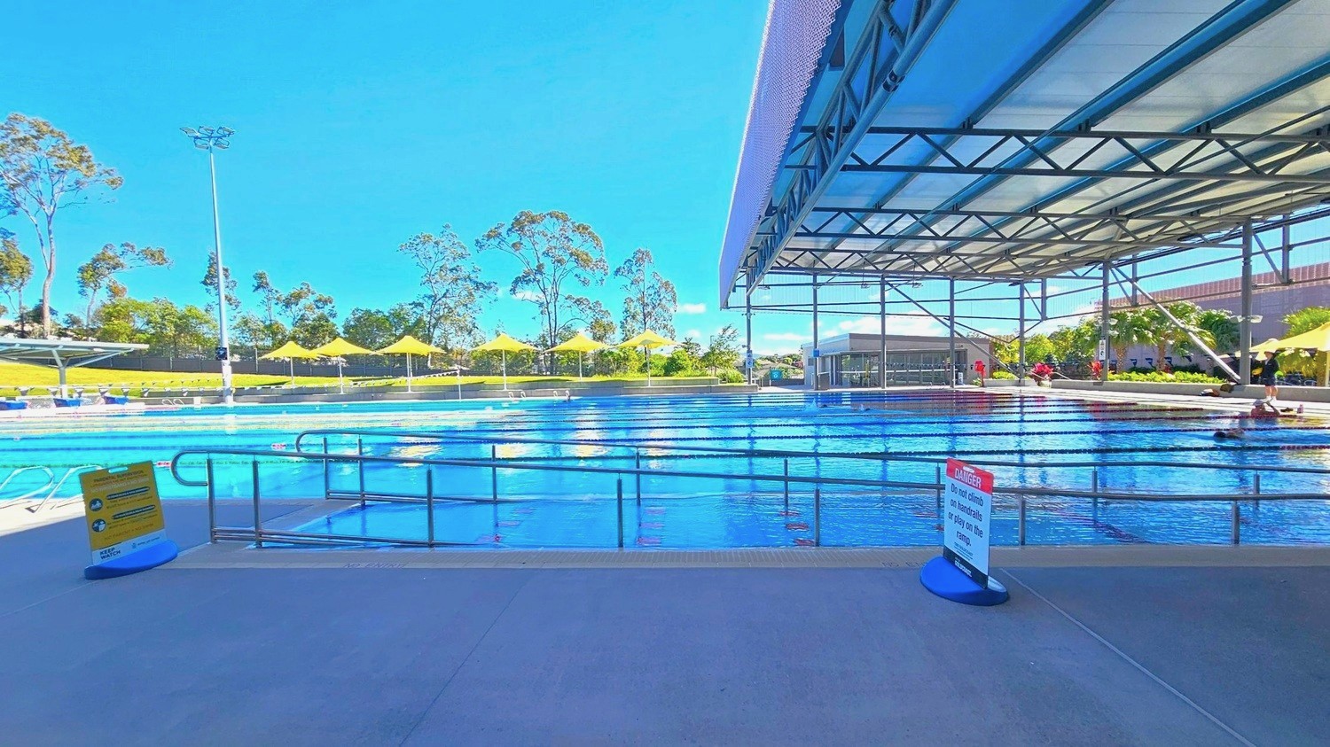 Outdoor swimming pool with multiple lanes, partially covered by a metal roof on the right. Yellow triangular shade structures line the poolside on the left. A sign in the foreground warns against climbing handrails. Trees and greenery are visible in the background, along with a tall light pole. The pool area is surrounded by a concrete deck.