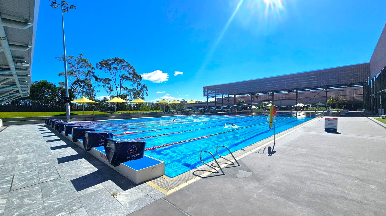 An outdoor swimming pool with multiple lanes, bordered by a tiled deck. On the left, a row of starting blocks and a tall light pole. In the background, yellow umbrellas and trees line the perimeter. On the right, a large shaded structure and a sign on the deck. The pool is bright blue, with swimmers in some lanes. The sky is clear and sunny.