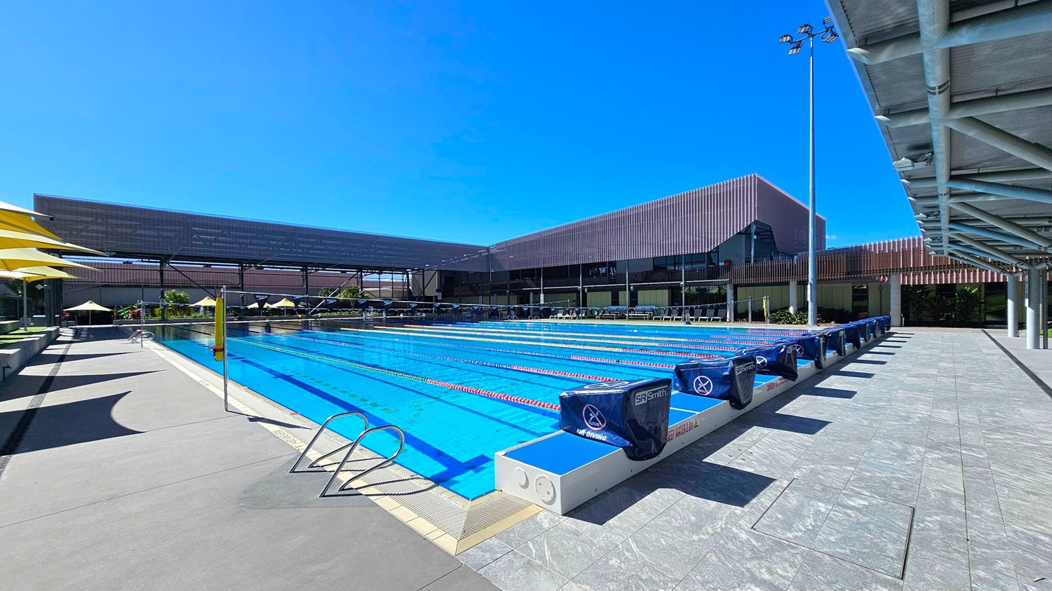 An outdoor swimming pool with multiple lanes, each marked by lane dividers. On the left, there are yellow umbrellas providing shade over seating areas. The pool deck is made of grey tiles. On the right, a covered walkway with a metal roof extends along the pool's edge. In the background, modern buildings with a mix of open and closed structures are visible, along with some greenery. The sky is clear and blue.