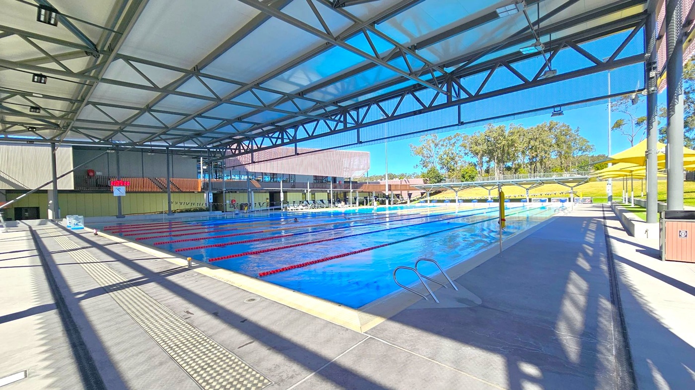 An outdoor swimming pool with multiple lanes, covered by a large metal and fabric structure. On the left, a digital clock is mounted on a wall. The pool is in the centre, with lane dividers and a ladder on the right. Yellow umbrellas and seating areas are visible on the right side. In the background, there are trees and a grassy area. The floor is concrete, and the ceiling is a metal framework with translucent panels.