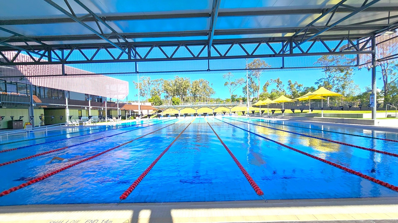 An outdoor swimming pool with multiple lanes, separated by red lane dividers, stretches into the distance. On the left, a building with large windows and a shaded area is visible. On the right, several yellow umbrellas provide shade over lounge chairs. In the background, trees and greenery are visible beyond the pool area. The ceiling is a metal structure with beams, partially covering the pool. The floor around the pool is tiled.