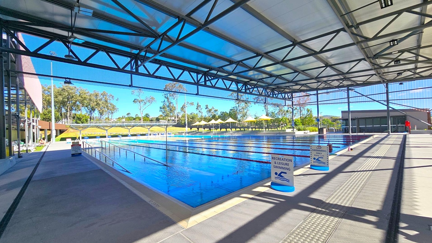 An indoor swimming pool with a high, metal-framed ceiling. The pool is divided into lanes, with signs indicating areas for recreation and lap swimming. On the left, there is a walkway with a few people sitting. The right side features a wide path with shadows from the ceiling structure. In the background, there are trees and yellow umbrellas providing shade over seating areas. The floor is tiled, and the ceiling has a transparent section allowing natural light in.
