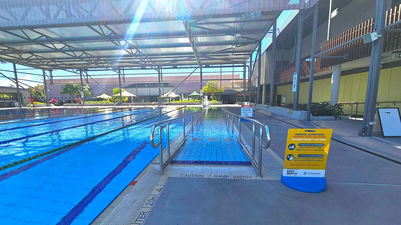 An indoor swimming pool with multiple lanes on the left, separated by lane dividers. In the centre, a ramp with metal railings leads into the water. On the right, a sign with safety instructions is placed on the pool deck. The ceiling is supported by metal beams, and the background features a building with large windows and some greenery, including palm trees. The floor is tiled near the pool and concrete on the deck.
