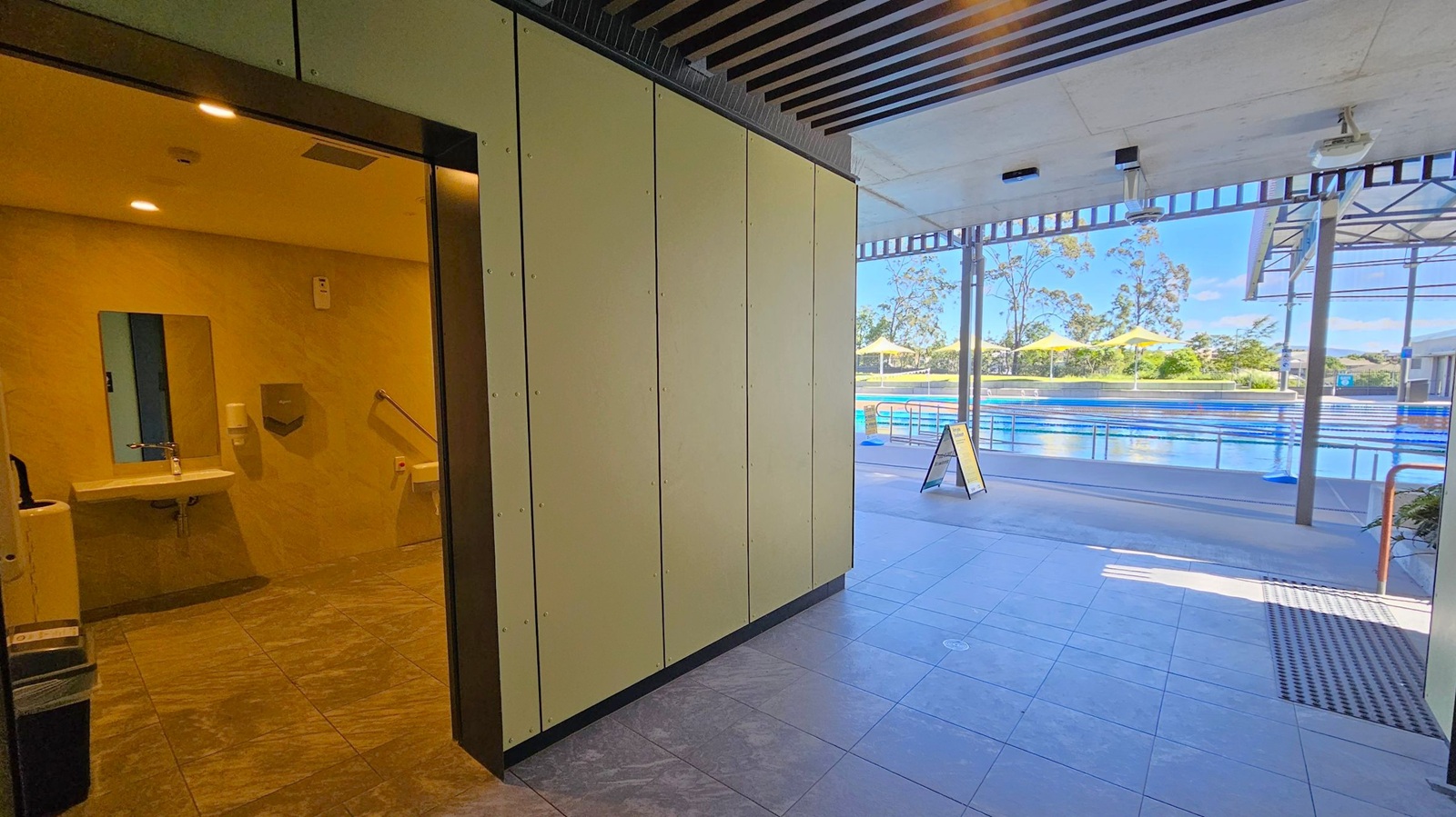 On the left, there's an entrance to a restroom with a visible sink, mirror, and handrails. The floor is tiled in a dark pattern. In the centre, a corridor leads to an outdoor area. On the right, there's a view of a swimming pool with lanes, surrounded by a deck. Yellow umbrellas and trees are visible in the background. The ceiling has a slatted design indoors and a flat design outdoors. A signboard stands near the pool area.