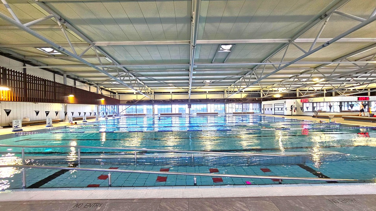 Indoor swimming pool with clear water and lane dividers. The ceiling features metal beams and panels. Large windows line the far wall, allowing natural light to illuminate the space. Digital clocks are mounted on the right wall, displaying the time. The pool deck is tiled, with a "NO ENTRY" sign visible. Safety railings run along the pool's edge. The left wall has vertical wooden slats and signage indicating swimming lanes. Indoor swimming pool with clear water and lane dividers. The ceiling features metal beams and panels. Large windows line the far wall, allowing natural light to illuminate the space. Digital clocks are mounted on the right wall, displaying the time. The pool deck is tiled, with a "NO ENTRY" sign visible. Safety railings run along the pool's edge. The left wall has vertical wooden slats and signage indicating swimming lanes.