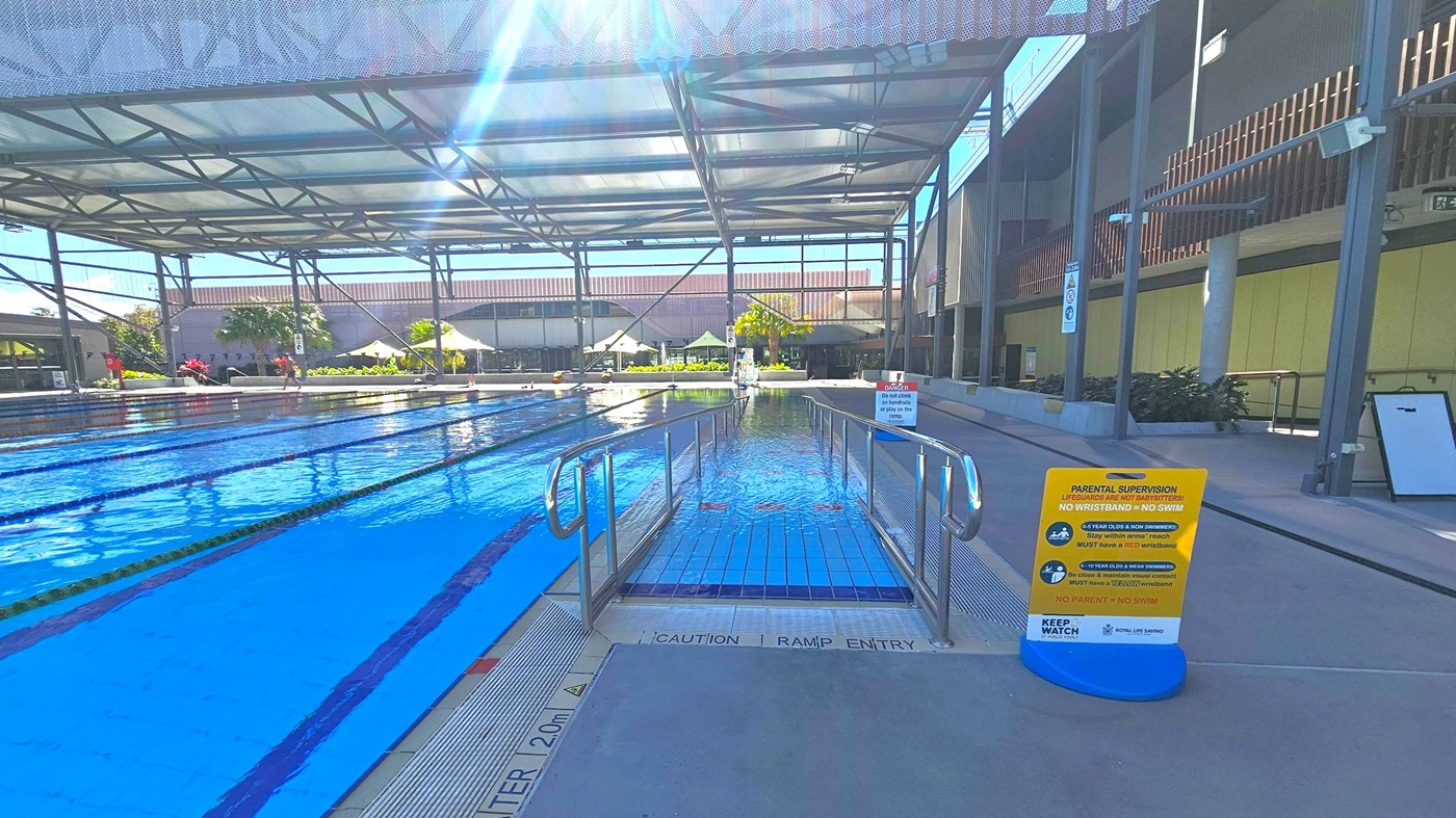 An indoor swimming pool with multiple lanes on the left, separated by lane dividers. In the centre, a ramp with metal railings leads into the water. On the right, a sign with safety instructions is placed on the pool deck. The ceiling is supported by a metal framework, and the background features a building with large windows and some greenery, including palm trees and plants. The floor is tiled near the pool and concrete on the deck. An indoor swimming pool with multiple lanes on the left, separated by lane dividers. In the centre, a ramp with metal railings leads into the water. On the right, a sign with safety instructions is placed on the pool deck. The ceiling is supported by a metal framework, and the background features a building with large windows and some greenery, including palm trees and plants. The floor is tiled near the pool and concrete on the deck.