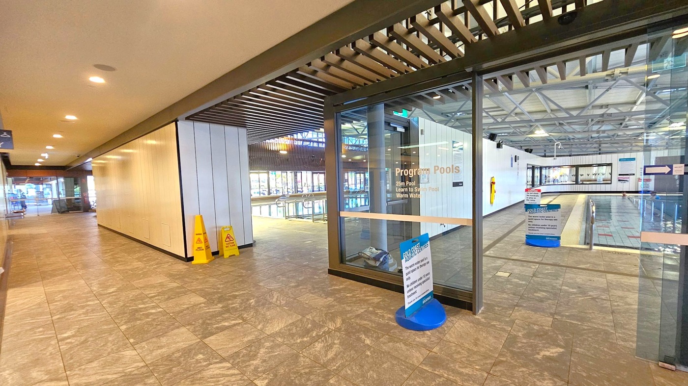 A hallway with a tiled floor and a ceiling with recessed lighting. On the right, there is a glass entrance to a pool area, labelled "Program Pools," with a view of the swimming pool inside. Two yellow caution signs are placed near the entrance. The left side of the hallway has a plain wall and leads to an open area with seating. The pool area has a high ceiling with exposed beams and large windows allowing natural light. A hallway with a tiled floor and a ceiling with recessed lighting. On the right, there is a glass entrance to a pool area, labelled "Program Pools," with a view of the swimming pool inside. Two yellow caution signs are placed near the entrance. The left side of the hallway has a plain wall and leads to an open area with seating. The pool area has a high ceiling with exposed beams and large windows allowing natural light.