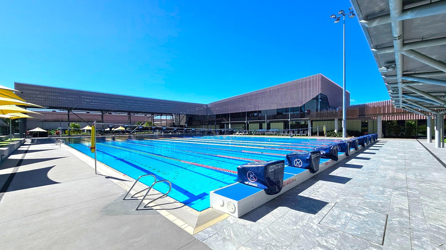 An outdoor swimming pool with multiple lanes, each marked by red and white lane dividers. On the left, there are yellow umbrellas providing shade over seating areas. The right side features a tiled walkway with a metal railing and a covered structure overhead. In the background, modern buildings with a mix of glass and metal elements are visible, along with more seating and umbrellas. The pool area is well-lit with tall floodlights. An outdoor swimming pool with multiple lanes, each marked by red and white lane dividers. On the left, there are yellow umbrellas providing shade over seating areas. The right side features a tiled walkway with a metal railing and a covered structure overhead. In the background, modern buildings with a mix of glass and metal elements are visible, along with more seating and umbrellas. The pool area is well-lit with tall floodlights.