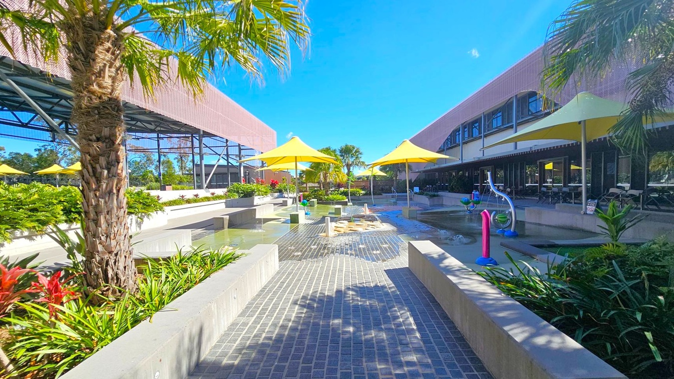 A courtyard with a paved walkway in the centre, flanked by low concrete walls and lush green plants. Yellow umbrellas provide shade over seating areas on both sides. On the left, a structure with a metal framework and a pink mesh roof is visible. On the right, a building with large windows and a shaded patio area. In the middle, water features and play equipment are present. Palm trees and other tropical plants are scattered throughout the area. A courtyard with a paved walkway in the centre, flanked by low concrete walls and lush green plants. Yellow umbrellas provide shade over seating areas on both sides. On the left, a structure with a metal framework and a pink mesh roof is visible. On the right, a building with large windows and a shaded patio area. In the middle, water features and play equipment are present. Palm trees and other tropical plants are scattered throughout the area.