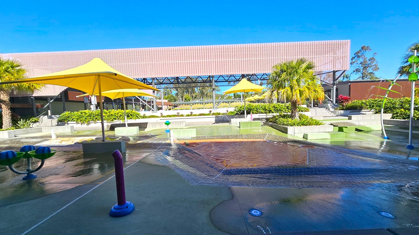A water play area with a wet surface and various water features, including a purple pipe and blue and green structures. Yellow umbrellas provide shade, and there are palm trees and lush greenery around. In the background, a large structure with a slatted roof and metal framework is visible, along with steps leading up to it. The area is surrounded by landscaped plants and shrubs. A water play area with a wet surface and various water features, including a purple pipe and blue and green structures. Yellow umbrellas provide shade, and there are palm trees and lush greenery around. In the background, a large structure with a slatted roof and metal framework is visible, along with steps leading up to it. The area is surrounded by landscaped plants and shrubs.