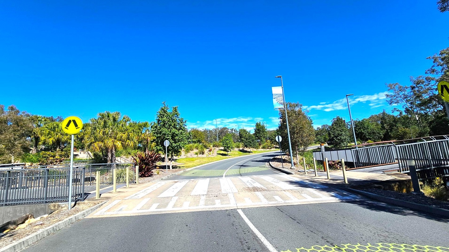 A road with a pedestrian crossing in the foreground, marked with white and green stripes. On the left, a yellow pedestrian sign is visible near a metal railing and lush greenery, including palm trees. On the right, a pathway with a metal fence runs parallel to the road. The background features a winding road lined with trees and street lamps, with a banner hanging from one of the poles. The scene is well-lit, suggesting a sunny day. A road with a pedestrian crossing in the foreground, marked with white and green stripes. On the left, a yellow pedestrian sign is visible near a metal railing and lush greenery, including palm trees. On the right, a pathway with a metal fence runs parallel to the road. The background features a winding road lined with trees and street lamps, with a banner hanging from one of the poles. The scene is well-lit, suggesting a sunny day.