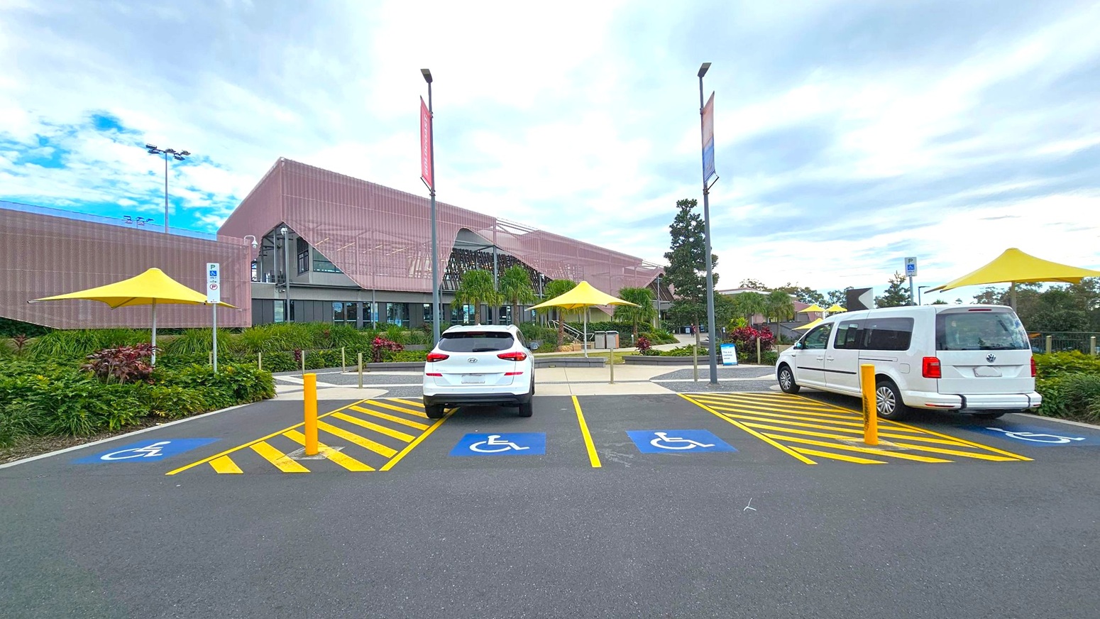 A modern building with a pink facade is in the background, featuring large glass windows and surrounded by greenery. In the foreground, there are two parked white vehicles in designated disabled parking spaces, marked with blue and yellow lines. Yellow umbrellas provide shade over nearby seating areas. Tall streetlights and banners line the pathway leading to the entrance. The ground is paved with asphalt. A modern building with a pink facade is in the background, featuring large glass windows and surrounded by greenery. In the foreground, there are two parked white vehicles in designated disabled parking spaces, marked with blue and yellow lines. Yellow umbrellas provide shade over nearby seating areas. Tall streetlights and banners line the pathway leading to the entrance. The ground is paved with asphalt.