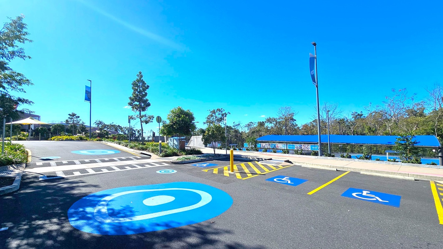 A parking area with designated disabled parking spaces marked in blue on the asphalt. On the left, there are trees and a pathway leading to a building with a white canopy. In the centre, a pedestrian crossing with white stripes is visible. On the right, a blue-roofed structure runs parallel to the parking spaces, with more trees and greenery in the background. Two tall street lamps with banners are positioned on either side of the parking area. A parking area with designated disabled parking spaces marked in blue on the asphalt. On the left, there are trees and a pathway leading to a building with a white canopy. In the centre, a pedestrian crossing with white stripes is visible. On the right, a blue-roofed structure runs parallel to the parking spaces, with more trees and greenery in the background. Two tall street lamps with banners are positioned on either side of the parking area.