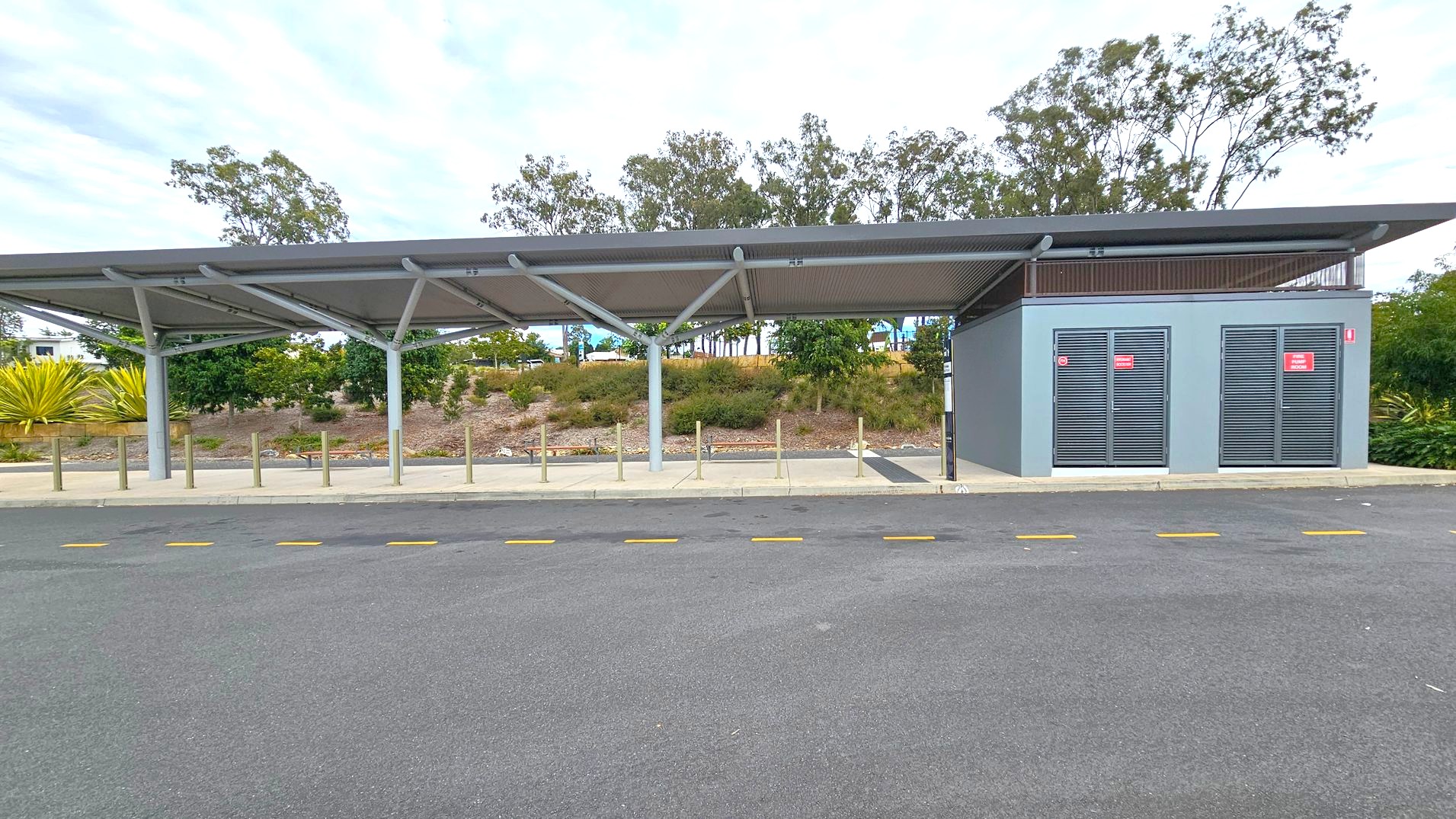 A bus stop with a modern shelter featuring a slanted roof supported by metal beams. On the right, there is a grey utility building with two vented doors displaying red signs. The foreground has a paved area with yellow dashed lines on the road. In the background, there are trees and shrubs on a slightly elevated grassy area. The sky is overcast. A bus stop with a modern shelter featuring a slanted roof supported by metal beams. On the right, there is a grey utility building with two vented doors displaying red signs. The foreground has a paved area with yellow dashed lines on the road. In the background, there are trees and shrubs on a slightly elevated grassy area. The sky is overcast.