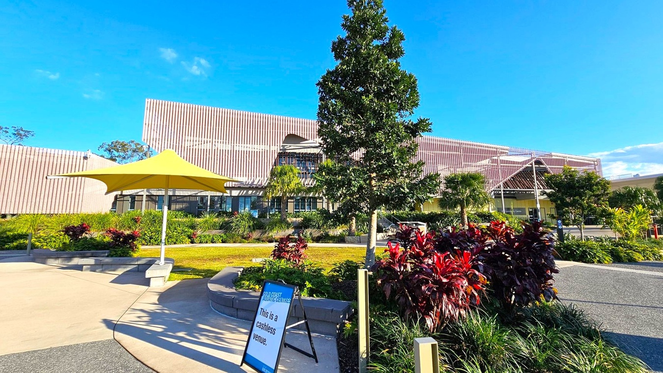 A modern building with a textured facade is in the background, surrounded by lush greenery and various plants. In the foreground, a yellow umbrella provides shade over a seating area. A sign on the left reads "This is a cashless venue." The ground is a mix of concrete and gravel paths, and a small tree is centrally located, adding to the landscaped environment. A modern building with a textured facade is in the background, surrounded by lush greenery and various plants. In the foreground, a yellow umbrella provides shade over a seating area. A sign on the left reads "This is a cashless venue." The ground is a mix of concrete and gravel paths, and a small tree is centrally located, adding to the landscaped environment.