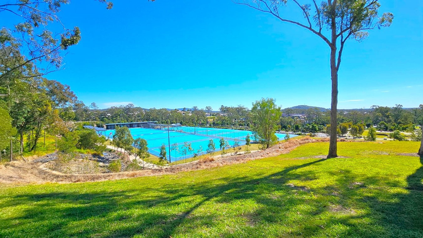 A grassy hill in the foreground with a tall tree on the right. In the middle, several bright blue tennis courts are visible, surrounded by a fence. To the left, there are more trees and a path. The background features more trees and a distant view of hills and buildings. A grassy hill in the foreground with a tall tree on the right. In the middle, several bright blue tennis courts are visible, surrounded by a fence. To the left, there are more trees and a path. The background features more trees and a distant view of hills and buildings.
