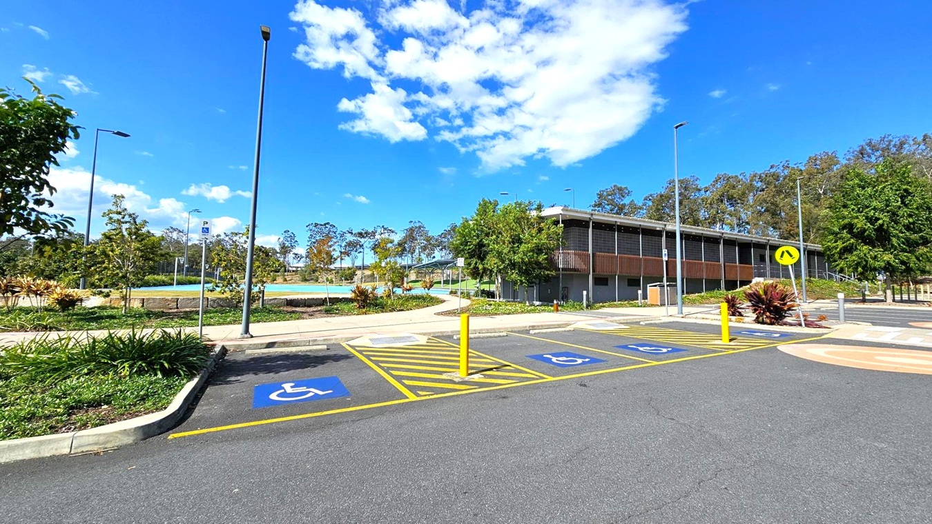 A parking area with designated disabled parking spaces marked in blue on the asphalt. Yellow lines and a bollard separate the spaces. On the right, a modern building with a flat roof and wooden slats is partially obscured by trees. In the background, a landscaped area with small trees and a pathway is visible. Several tall streetlights are scattered throughout the scene. A parking area with designated disabled parking spaces marked in blue on the asphalt. Yellow lines and a bollard separate the spaces. On the right, a modern building with a flat roof and wooden slats is partially obscured by trees. In the background, a landscaped area with small trees and a pathway is visible. Several tall streetlights are scattered throughout the scene.