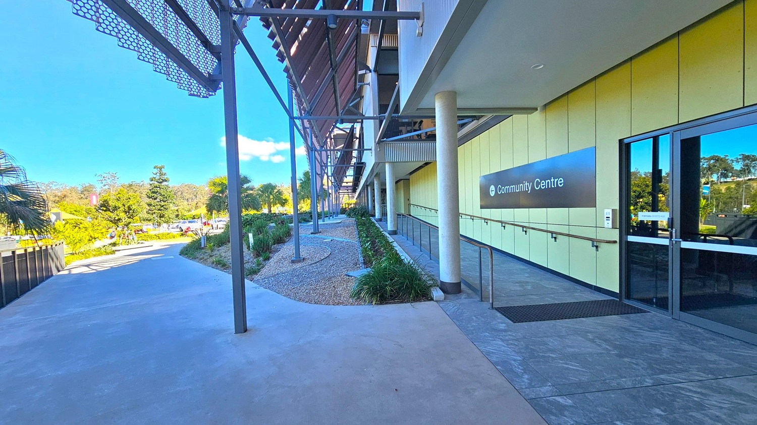 A pathway leads to a modern building on the right, marked as a "Community Centre" with a sign. The building features a glass entrance and a series of vertical columns. On the left, there is a landscaped area with small plants and trees, bordered by a metal fence. Overhead, a metal awning with a geometric pattern provides shade. The ground is paved with concrete, and the area is well-lit by natural light. A pathway leads to a modern building on the right, marked as a "Community Centre" with a sign. The building features a glass entrance and a series of vertical columns. On the left, there is a landscaped area with small plants and trees, bordered by a metal fence. Overhead, a metal awning with a geometric pattern provides shade. The ground is paved with concrete, and the area is well-lit by natural light.