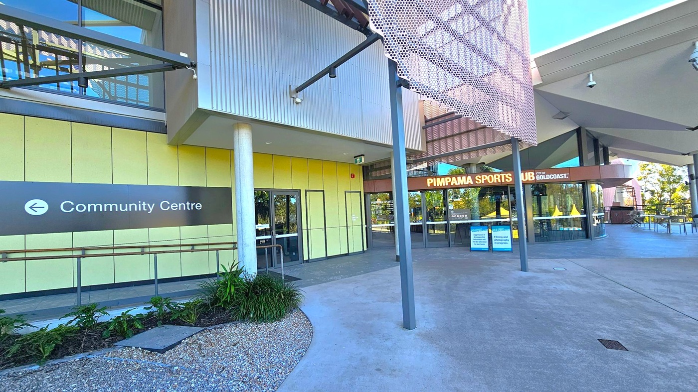 Modern building exterior with a sign pointing left to the "Community Centre" on the left side. The wall is clad in yellow panels with a glass door entrance. In the centre, a cylindrical column supports the structure. On the right, the entrance to "Pimpama Sports Hub" is visible, with large glass windows and a sign above. The foreground features a concrete path and landscaped area with small plants and gravel. The ceiling has a geometric design with metal and glass elements. Modern building exterior with a sign pointing left to the "Community Centre" on the left side. The wall is clad in yellow panels with a glass door entrance. In the centre, a cylindrical column supports the structure. On the right, the entrance to "Pimpama Sports Hub" is visible, with large glass windows and a sign above. The foreground features a concrete path and landscaped area with small plants and gravel. The ceiling has a geometric design with metal and glass elements.