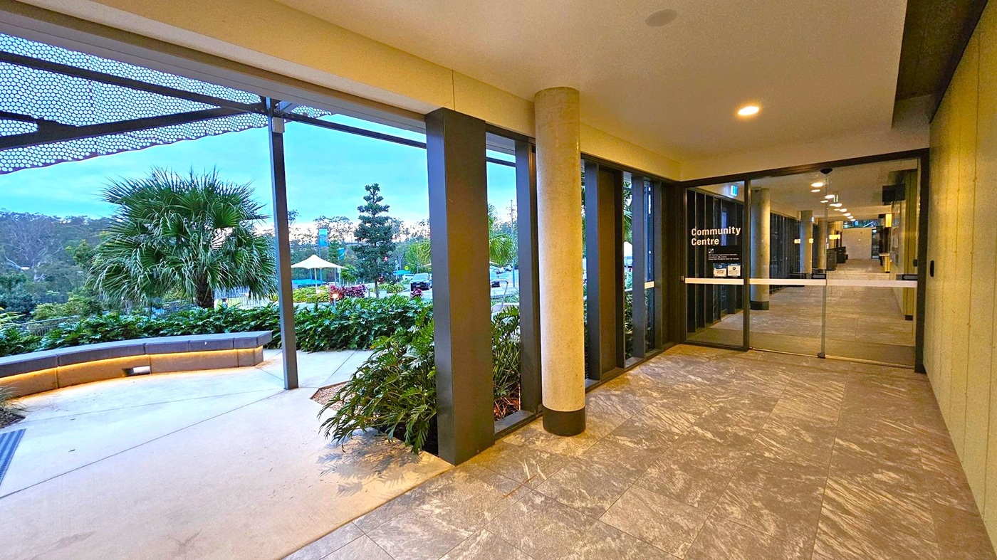 A corridor with a tiled floor leads to a glass door marked "Community Centre" on the right. The ceiling has recessed lighting. On the left, large windows reveal an outdoor area with a curved bench, lush greenery, and a palm tree. A metal canopy with a perforated design extends overhead. In the background, there are more trees and a structure with a yellow canopy. A corridor with a tiled floor leads to a glass door marked "Community Centre" on the right. The ceiling has recessed lighting. On the left, large windows reveal an outdoor area with a curved bench, lush greenery, and a palm tree. A metal canopy with a perforated design extends overhead. In the background, there are more trees and a structure with a yellow canopy.